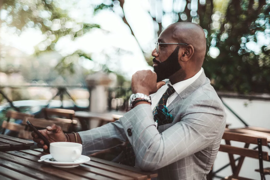 Dark skinned man with immaculately groomed beard, wearing an expensive business suit and watch, sits at an outdoor cafe.