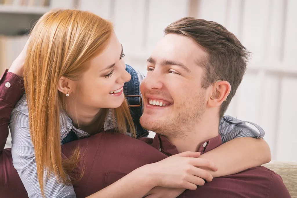 A young man with hearing loss wearing a Bluetooth hearing aid lovingly hugs a young women